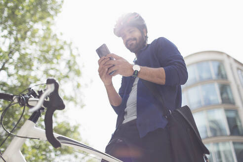 Mann benutzt Smartphone auf Fahrrad unter sonnigem Gebäude - CAIF29680