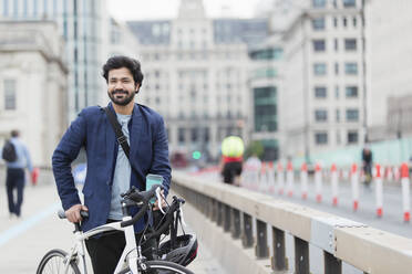 Porträt lächelnder Geschäftsmann mit Fahrrad und Kaffee auf einer Stadtbrücke - CAIF29686