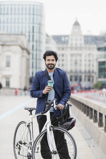Porträt lächelnder Geschäftsmann mit Kaffee und Fahrrad auf einer Stadtbrücke - CAIF29691