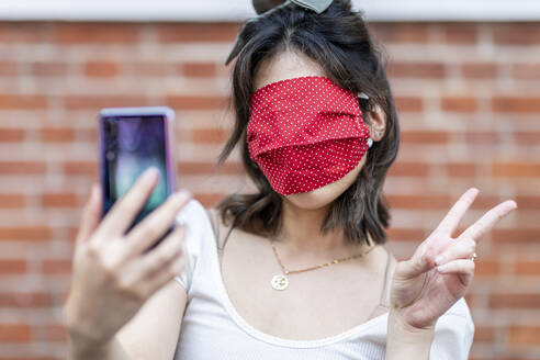 Nahaufnahme eines mit einer Maske bedeckten Frauengesichts, das ein Selfie vor einer Backsteinmauer macht - WPEF03345