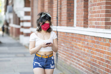 Young woman's face covered with mask using smart phone while standing in city - WPEF03346