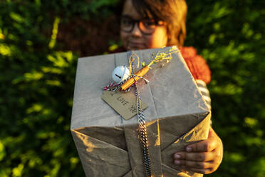 Nahaufnahme eines Jungen, der ein Weihnachtsgeschenk in der Hand hält, während er gegen Pflanzen im Garten steht - VABF03457