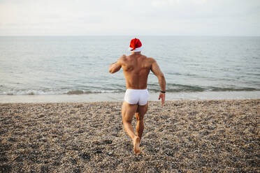 Shirtless young man wearing Santa hat walking at beach during sunset - MIMFF00222