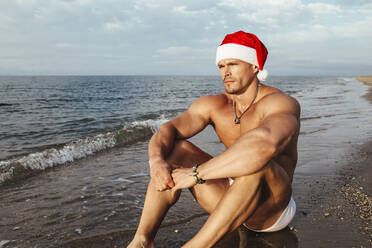 Nachdenklicher junger Mann ohne Hemd mit Weihnachtsmannmütze am Strand sitzend - MIMFF00225