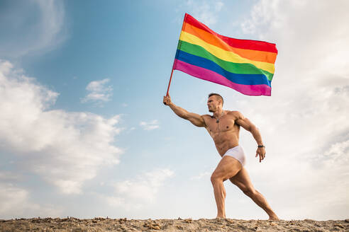 Hemdloser junger Mann mit Regenbogenflagge, der am Strand gegen den Himmel läuft - MIMFF00227