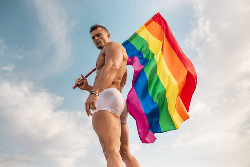 Hemdloser junger Mann hält Regenbogenflagge, während er gegen den Himmel steht - MIMFF00228
