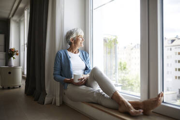 Lächelnde ältere Frau, die durch das Fenster schaut, während sie zu Hause sitzt - RBF07784