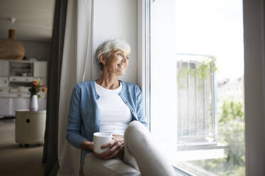 Ältere Frau schaut aus dem Fenster, während sie zu Hause sitzt - RBF07785
