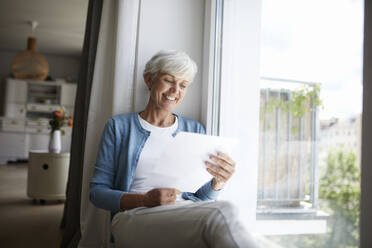 Senior woman reading letter happily while sitting on window at home - RBF07790