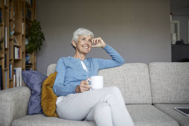 Nachdenkliche ältere Frau, die zu Hause auf dem Sofa sitzt und eine Kaffeetasse hält - RBF07793