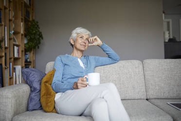 Nachdenkliche ältere Frau, die zu Hause auf dem Sofa sitzt und eine Kaffeetasse hält - RBF07794
