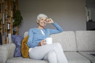 Lächelnde ältere Frau sitzt auf dem Sofa und hält eine Kaffeetasse in der Hand - RBF07795