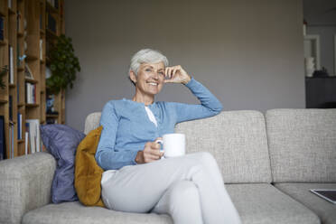 Smiling senior woman sitting on sofa holding coffee cup at home - RBF07795