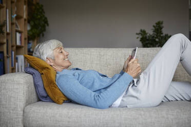 Lächelnde Frau, die ein digitales Tablet benutzt, während sie zu Hause auf dem Sofa liegt - RBF07803