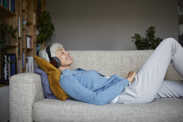 Senior woman listening to music while lying down on sofa at home - RBF07804