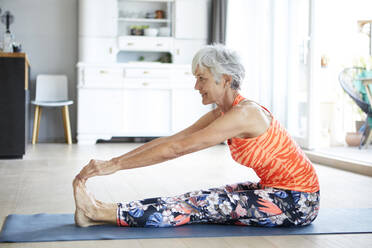 Fitte ältere Frau, die ihre Beine und ihren Rücken dehnt, während sie zu Hause trainiert - RBF07807