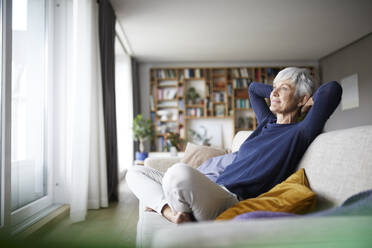 Ältere Frau mit Händen hinter dem Kopf mit gekreuzten Beinen auf dem Sofa zu Hause sitzen - RBF07811