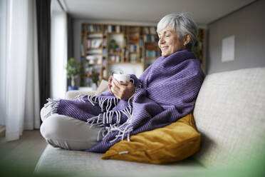 Thoughtful senior woman with blanket holding coffee cup while sitting on sofa at home - RBF07812