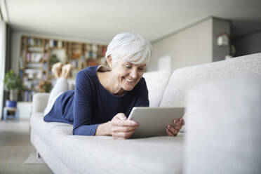 Senior woman using digital tablet lying on sofa at home - RBF07814