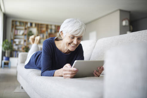 Ältere Frau mit digitalem Tablet zu Hause auf dem Sofa liegend - RBF07814