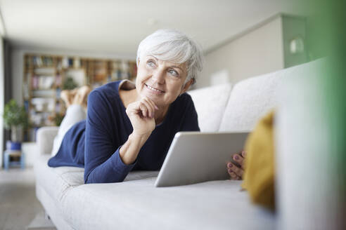 Ältere Frau mit Hand am Kinn, die ein digitales Tablet benutzt, während sie zu Hause auf dem Sofa liegt - RBF07815