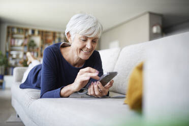 Ältere Frau benutzt ein Smartphone, während sie zu Hause auf dem Sofa liegt - RBF07816