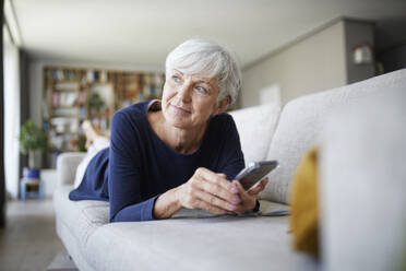 Aktive ältere Frau, die ein Smartphone benutzt und wegschaut, während sie zu Hause auf dem Sofa liegt - RBF07817