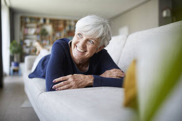 Lächelnde ältere Frau, die mit verschränkten Armen wegschaut, während sie zu Hause auf dem Sofa liegt - RBF07820