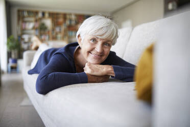 Lächelnde ältere Frau mit Hand am Kinn auf dem Sofa zu Hause liegend - RBF07821