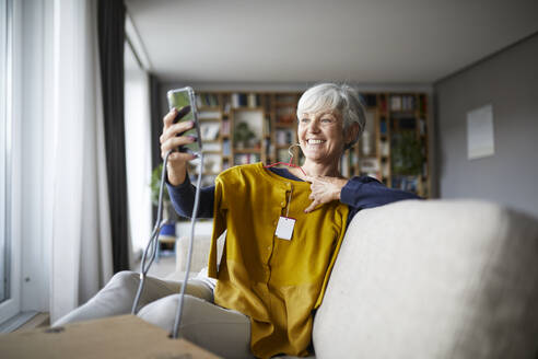Smiling senior woman taking selfie with new blouse on smart phone while sitting at home - RBF07824