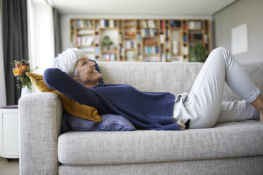 Smiling senior woman with hands behind head lying down on sofa at home - RBF07826