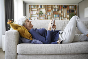 Ältere Frau schreibt Textnachrichten auf einem digitalen Tablet, während sie zu Hause auf dem Sofa liegt - RBF07827