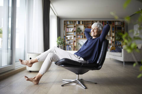 Happy senior woman with hands behind head sitting on chair at home stock photo