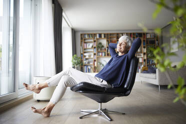 Nachdenkliche ältere Frau mit Händen hinter dem Kopf, die zu Hause auf einem Stuhl sitzt - RBF07830