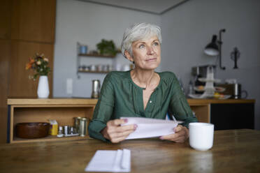 Ältere Frau, die einen Brief liest und wegschaut, während sie zu Hause sitzt - RBF07833