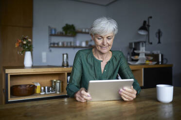 Ältere Frau, die zu Hause sitzend in ein digitales Tablet schaut - RBF07834