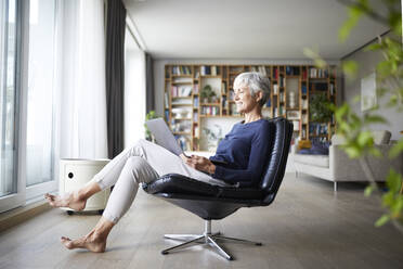 Aktive ältere Frau, die einen Laptop benutzt, während sie zu Hause auf einem Stuhl sitzt - RBF07844