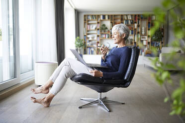 Active senior woman sending voicemail on smart phone while using laptop sitting at phone - RBF07845