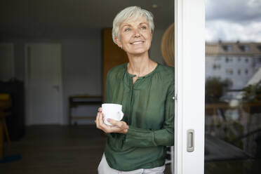 Ältere Frau steht mit Kaffeetasse am Fenster zu Hause - RBF07860