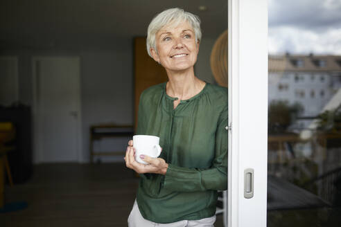 Ältere Frau steht mit Kaffeetasse am Fenster zu Hause - RBF07860