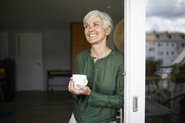 Senior woman looking away standing with coffee cup by window at home - RBF07861