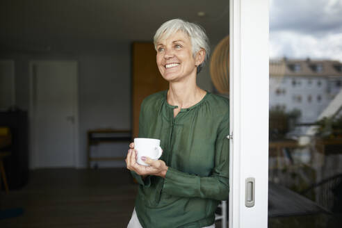 Ältere Frau, die mit einer Kaffeetasse am Fenster steht und wegschaut, zu Hause - RBF07861
