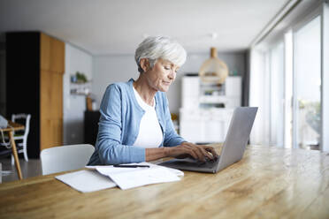 Ältere Frau arbeitet am Laptop, während sie zu Hause auf einem Stuhl sitzt - RBF07865