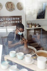 Männlicher Töpfer mit Maske, der in einer Werkstatt durch ein Fenster gesehen Ton in Form bringt - EGAF00711