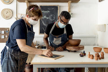 Coworkers wearing masks discussing designs over slate on table in workshop - EGAF00732
