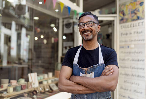 Confident male potter with arms crossed standing outside ceramic shop - EGAF00734