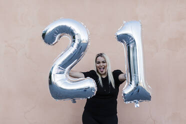 Fröhliche junge Frau hält Luftballons mit der Nummer 21, während sie an der Wand steht - DCRF00840
