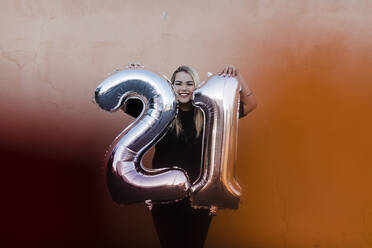 Glückliche junge Frau hält Luftballons mit der Nummer 21, während sie an der Wand steht - DCRF00842