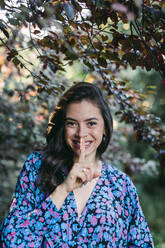 Close-up of smiling young woman with finger on lips standing against plants in park - DCRF00853