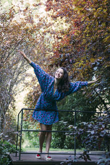 Fröhliche junge Frau mit ausgestreckten Armen, die an einem Geländer gegen Bäume in einem Park steht - DCRF00854